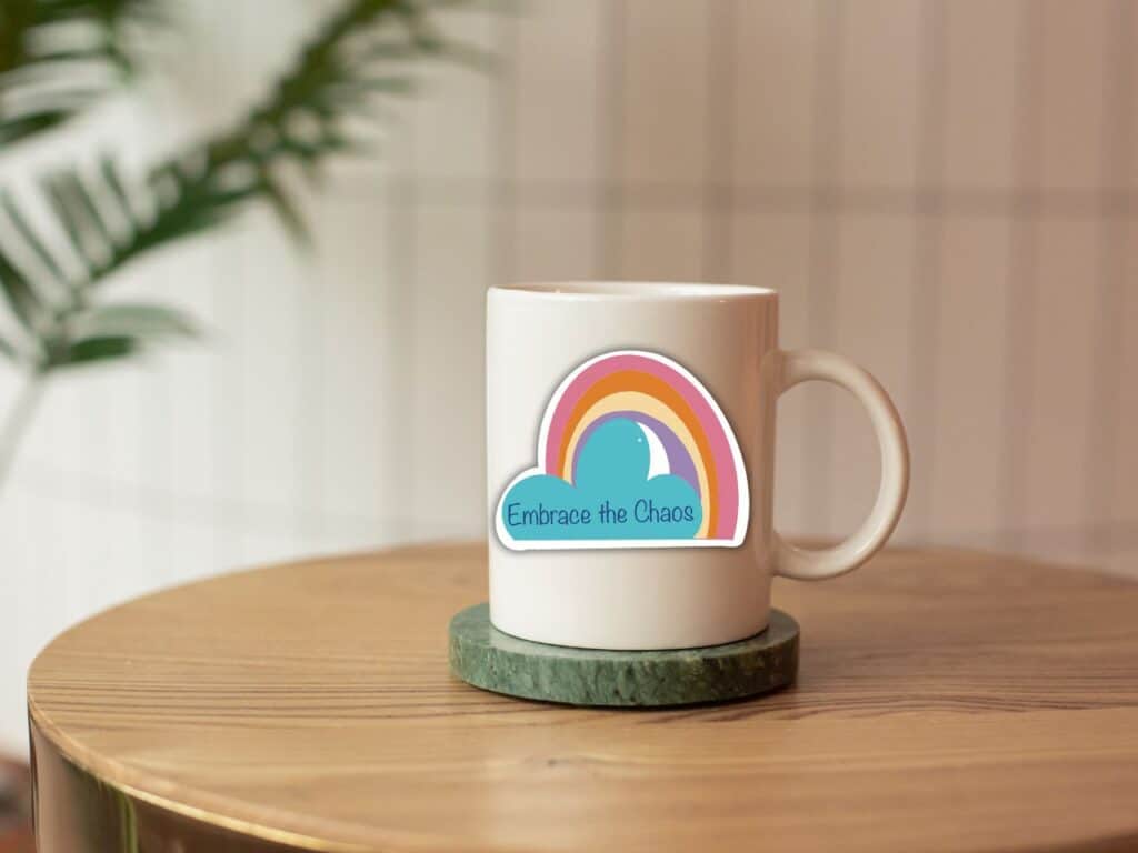 coffee cup with embrace the chaos rainbow and cloud sticker, sitting on a wooden table