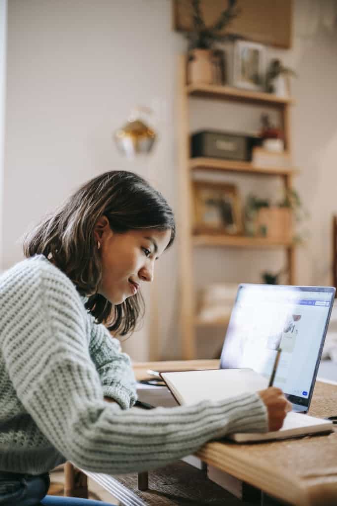 Side view of smart focused female writing in notebook while using netbook at table on blurred background