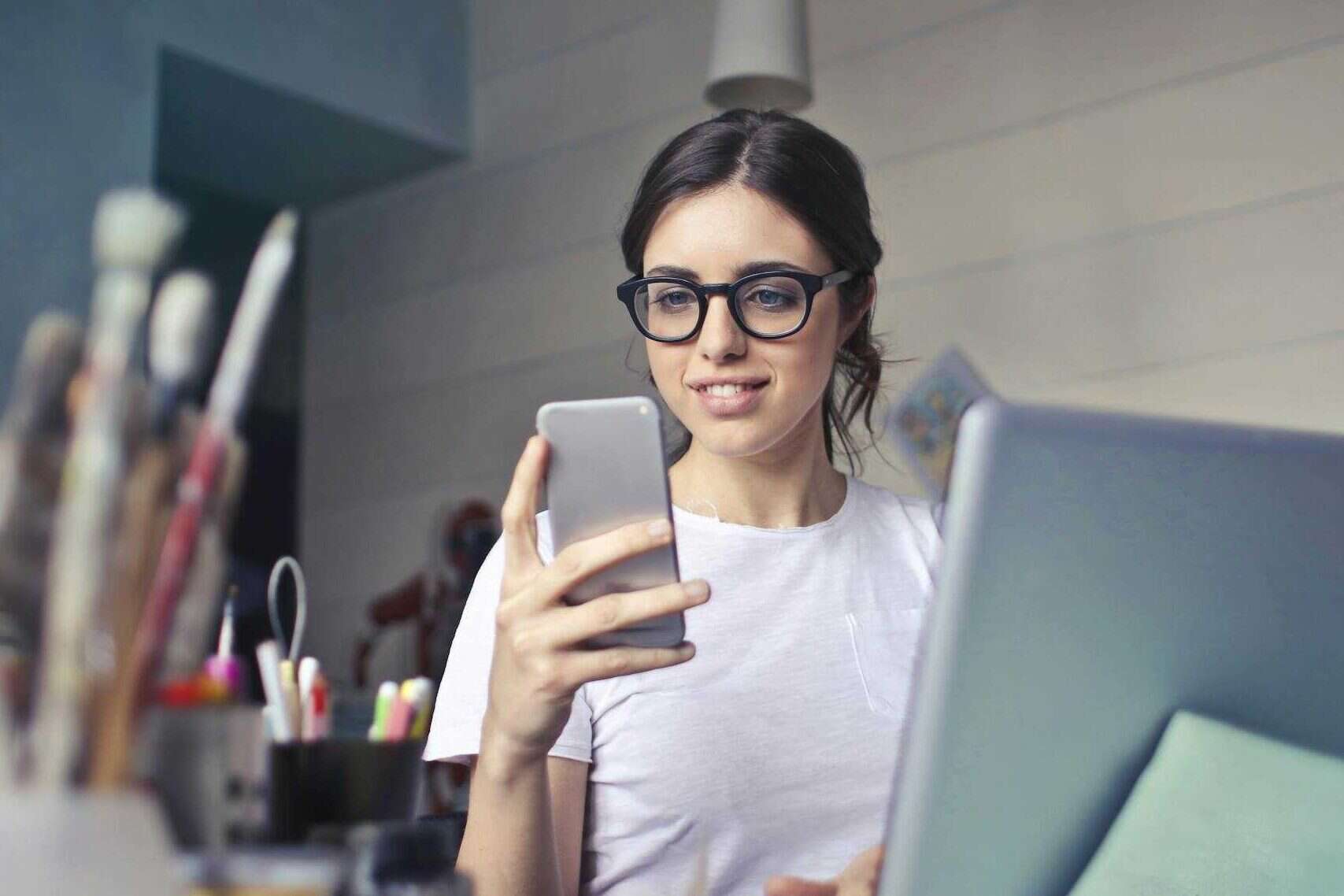 A woman using her phone at a desk, surrounded by art supplies and a laptop, in a creative workspace selling digital art