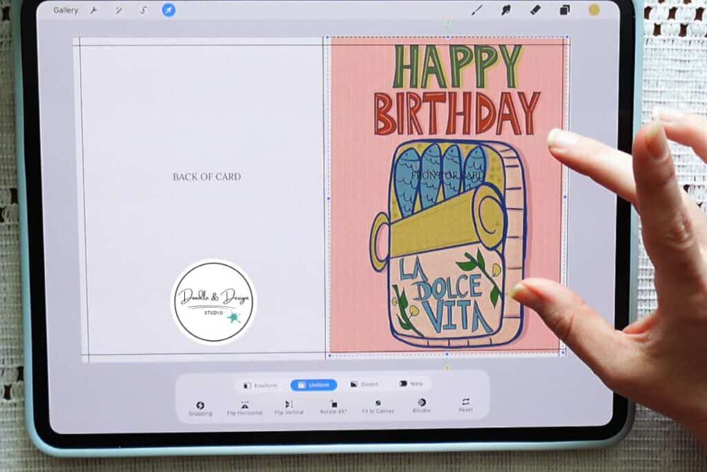 A hand adjusts a digital birthday card design on a tablet while creating printable greeting card artwork.