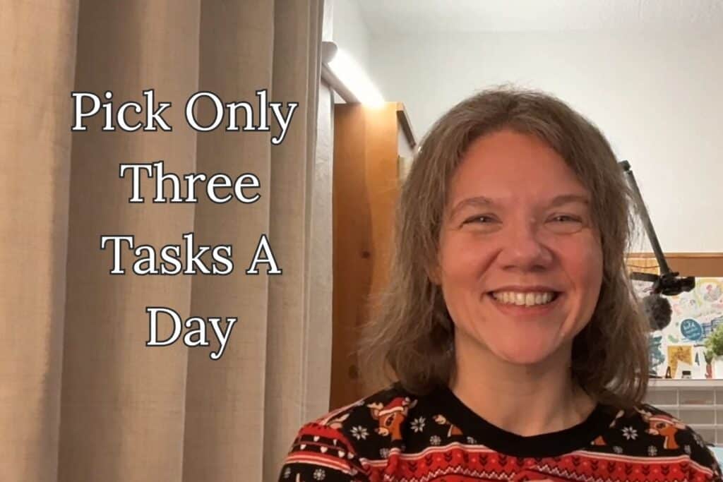 A woman smiles in front of a curtain and office setup, with the phrase “Pick Only Three Tasks A Day” encouraging a simplified task approach.
