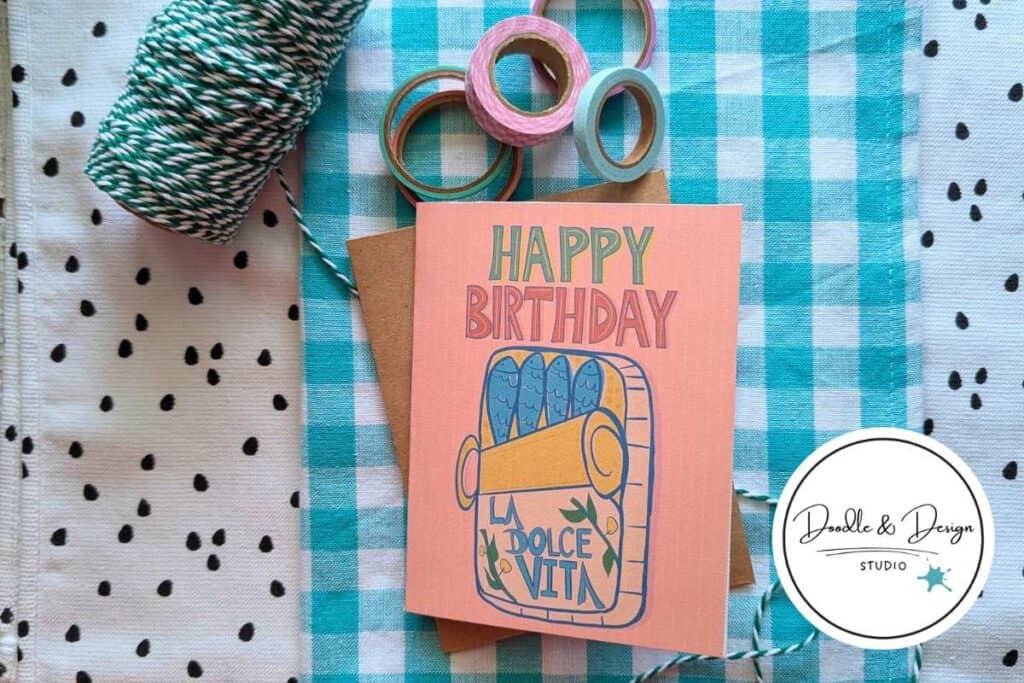 A handmade illustrated birthday card with a sardine tin design sits on a teal gingham cloth surrounded by twine, washi tape, and craft supplies.